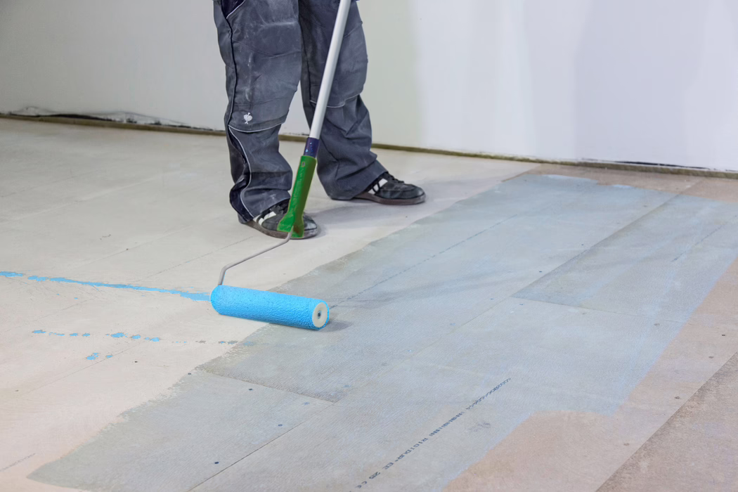 Si vedono gli stinchi e i piedi di un operaio mentre con un rullo a manico lungo applica un primer o una vernice azzurro chiaro su un sottofondo in legno. La persona indossa pantaloni da lavoro scuri e scarpe da ginnastica nere; il rullo lascia uno strato blu bagnato sulle tavole del pavimento in una stanza con pareti bianche. L'immagine mette in evidenza la preparazione del pavimento, gli strumenti e le macchie di vernice.