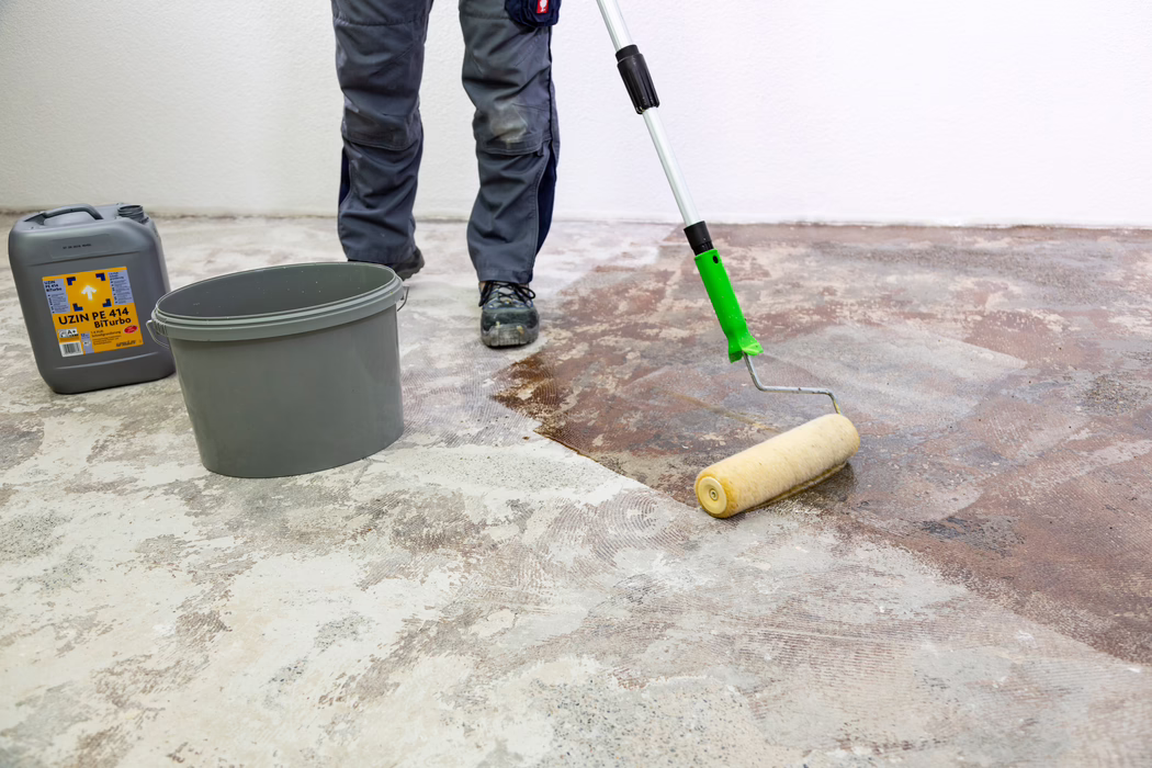 UZIN_Application Image_PE 414_9406_2018-11 Sono visibili gli stinchi e le scarpe di un operaio mentre con un rullo a manico lungo applica un primer o adesivo di colore marrone su un pavimento in cemento nudo. Vicino, contro una parete bianca, ci sono un secchio di plastica grigio e un contenitore grigio con l'etichetta "UZIN PE 414 Biturbo". L'operaio indossa pantaloni da lavoro grigi e scarpe antinfortunistiche nere; la superficie del pavimento mostra residui di vecchi adesivi e il calcestruzzo levigato mentre il rullo stende il prodotto in modo uniforme per preparare il sottofondo.